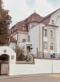 White house with brown roof, white walls, several windows, fenced off with white wall