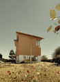 Two-story wooden house with large windows, surrounded by a field of wildflowers and trees. The house appears to be situated in a rural or natural setting with a scenic view.