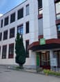 A multi-story commercial or office building with a modern, gray and red exterior design. The building has numerous windows, a prominent entrance marked with the number '44', and landscaping elements like a tree and flowers around the entrance.