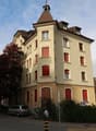 3 story building, beige exterior, red shutters, tower, cars parked in front