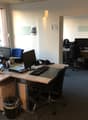 The image shows an office space with multiple desks, computers, and office equipment. The room has large windows providing natural light, and the walls are painted a light color. There are various office supplies and documents visible on the desks.