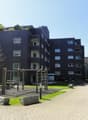A multi-story apartment building with a modern, brick facade. The building has balconies on several floors and is surrounded by a landscaped courtyard with paved walkways.