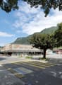 The image shows a street view of a town or city. There is a large supermarket building with the Migros logo visible. The street has a pedestrian crossing and parked cars. The surrounding area is lush with trees and greenery, and there are mountains visibl
