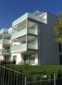 3 story apartment building with balconies, white exterior with glass railings, fenced yard, lawn, and trees