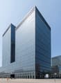 Large modern glass and steel office building with a rectangular, geometric design. The building has multiple floors and a prominent facade with a grid-like pattern of windows. There are trees and other buildings visible in the surrounding area.
