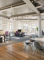 Large open-plan office space with high ceilings, large windows, and modern furniture. The space features workstations with computers, conference tables, and a mix of seating options. The decor is minimalist with a neutral color palette and wooden flooring