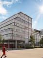 An artist's impression of a modern, multi-story building with raised ground floor, surrounded by trees and parked vehicles.
