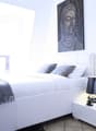 white bed, white nightstand, white lamp, white orchid, gray blanket, gray pillows, white walls, white ceiling, white floor, painting of Buddha