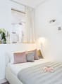 Modern small bedroom with tiled floor, a double bed with a gray and white striped bedspread, a nightstand with magazines, built-in wardrobe, a curtain for the shower, shelves with decorative items, a plant, and a decorative lamp.