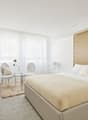 Modern bedroom with beige bed, white headboard, two white armchairs, round glass table, decorative rug, large windows with white curtains, and a desk with a television on the wall.