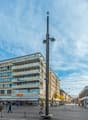 The image shows a busy city street with a mix of commercial and residential buildings. There is a tall lamp post with multiple cameras mounted on it in the center of the frame. People can be seen walking along the sidewalks and crossing the street. The bu