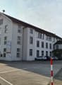 3 story building, white exterior, multiple windows, brown roof, parking lot, cars parked