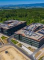 Large modern office complex with multiple interconnected buildings, surrounded by green landscapes and mountains in the background. The buildings have a sleek, contemporary design with glass facades and solar panels on the roofs. There is a parking lot in