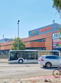 A large Migros supermarket building with a bus stop and people riding bicycles in front of it. The building has a distinctive orange and blue color scheme.