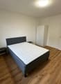 The image shows a bedroom with a double bed, a bedside table, and a wardrobe. The room has white walls and hardwood flooring.