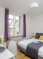 Spacious bedroom with large windows, gray upholstered bed, white bedding, purple curtains, and a vase of yellow tulips on a side table