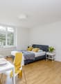 Bright and spacious studio apartment with large windows, hardwood floors, a double bed, a dining table, and a TV stand. The room has a cozy and modern feel with yellow accents and decorative elements.