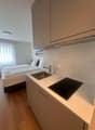 The image shows a studio apartment with a kitchenette area. The kitchenette has a sink, a cooktop, and a dishwasher. There is a bed visible in the background, indicating this is a studio apartment with a combined living and sleeping space.