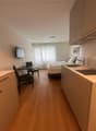 Compact studio apartment with hardwood floors, white walls, a bed, a desk with a chair, and kitchen cabinets with a dishwasher visible.