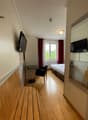 The image shows a bedroom with a bed, a desk, and two TVs mounted on the walls. The room has hardwood floors and large windows with red curtains. The overall decor appears to be modern and minimalist.