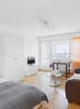 Bright and spacious studio apartment with large windows offering a view of the city. The room features a bed, a sofa, a dining table, and a TV stand. The walls are decorated with a colorful landscape painting. The apartment has hardwood floors and modern