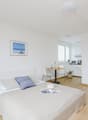 white walls, wooden floor, single bed, pillows, hat, table with flowers, kitchenette, refrigerator, picture frames