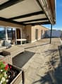 A covered outdoor terrace with a view, featuring a patio furniture set, a table, a large flower pot with flowers, and a storage box on the right side.