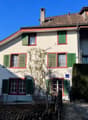 White house with green shutters and red trims, brown roof, several windows, small tree in front, trash bins beside the entrance