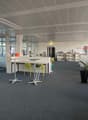 The image shows a modern and open office space with a high ceiling, glass walls, and a mix of workstations and lounge areas. The space has a gray carpet floor, white walls, and a gray ceiling with exposed beams. There are several desks with chairs, shelve