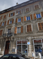 Multi-story building with a traditional European architectural style, featuring stone walls, wooden shutters, and a balcony. The building houses various commercial establishments, including a lottery shop, an art studio, and a cafeteria.