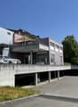 A multi-story commercial or residential building with a parking area underneath. The building has a modern, angular design with a mix of materials including concrete, metal, and glass. There are several cars parked in the covered parking area below the bu