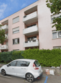 3-story apartment building with balconies, surrounded by trees and greenery, with parking spaces in front