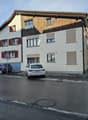 3 story house, white walls, brown roof, red shutters, cars parked