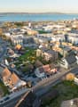 Aerial view of a city with a mix of residential and commercial buildings, surrounded by a body of water and hills in the background. The city appears to have a mix of older and newer buildings, with a variety of architectural styles. There are roads, park