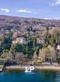 Aerial view of a hillside town with several houses, surrounded by trees and mountains, with a shoreline and boats