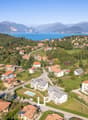 Numerous residential buildings, some with solar panels, surrounded by lush greenery, multiple houses with balconies, a swimming pool in front of a building, mountainous terrain, a large body of water in the background, and a clear blue sky.