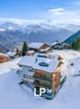Snow-covered mountain village with wooden chalets, apartment buildings, and scenic mountain views in the background