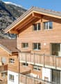 A multi-story wooden chalet-style building with a sloped roof and balconies overlooking a snowy mountain landscape. The building has large windows and a wooden exterior. There are other similar buildings and a village visible in the background.