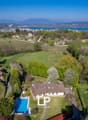 Aerial view of a large house with a swimming pool surrounded by lush greenery, with a scenic view of a lake and mountains in the background.