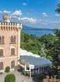 A large, pink-colored castle with turrets and arched windows, surrounded by lush greenery and overlooking a body of water in the distance.