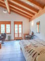 Spacious bedroom with wooden beamed ceiling, large windows providing natural light, tile flooring, leather armchair, desk, and a patterned bedspread on the bed.