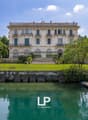 A large, multi-story historic building with a classical architectural style, surrounded by lush greenery and a body of water. The building has balconies, arched windows, and a decorative roofline.