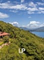 A large, two-story house with a red roof situated on a hillside overlooking a lake and mountains. The house is surrounded by lush, green vegetation and trees. The landscape features a scenic view of the lake and mountains in the distance.