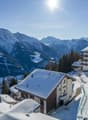 A group of snow-covered houses on a hillside with a ski track, surrounded by mountains and trees. The sun is shining brightly in the sky.
