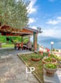 Garden with covered terrace, lake view, stairs to balcony, wooden patio table, chairs, greenery, flowers, olive tree, mountain background