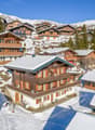 Mountain village, multiple wooden chalets, snow-covered rooftops, surrounded by trees and mountains