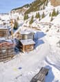 Wooden houses, snow covered mountain, ski lift in the background, trees and shrubs, a house under construction