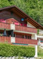 Apartment building, multiple wooden balconies, red shutters, hedge, grassy hillside, stone retaining wall
