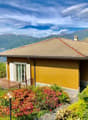 Two-story house with a yellow exterior, a tiled roof, and a balcony overlooking a scenic lake and mountains. The house is surrounded by lush greenery and colorful flowers in the garden.