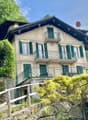 2-story house with a traditional Alpine-style architecture, featuring a sloped roof, wooden balconies, and green shutters. The house is surrounded by lush greenery and situated on a hillside overlooking a scenic landscape.